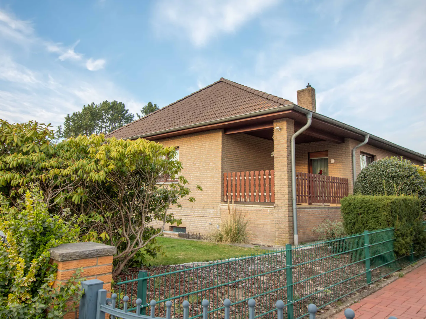 A single-story brick house with a brown roof, a small porch, and a green metal fence in front.