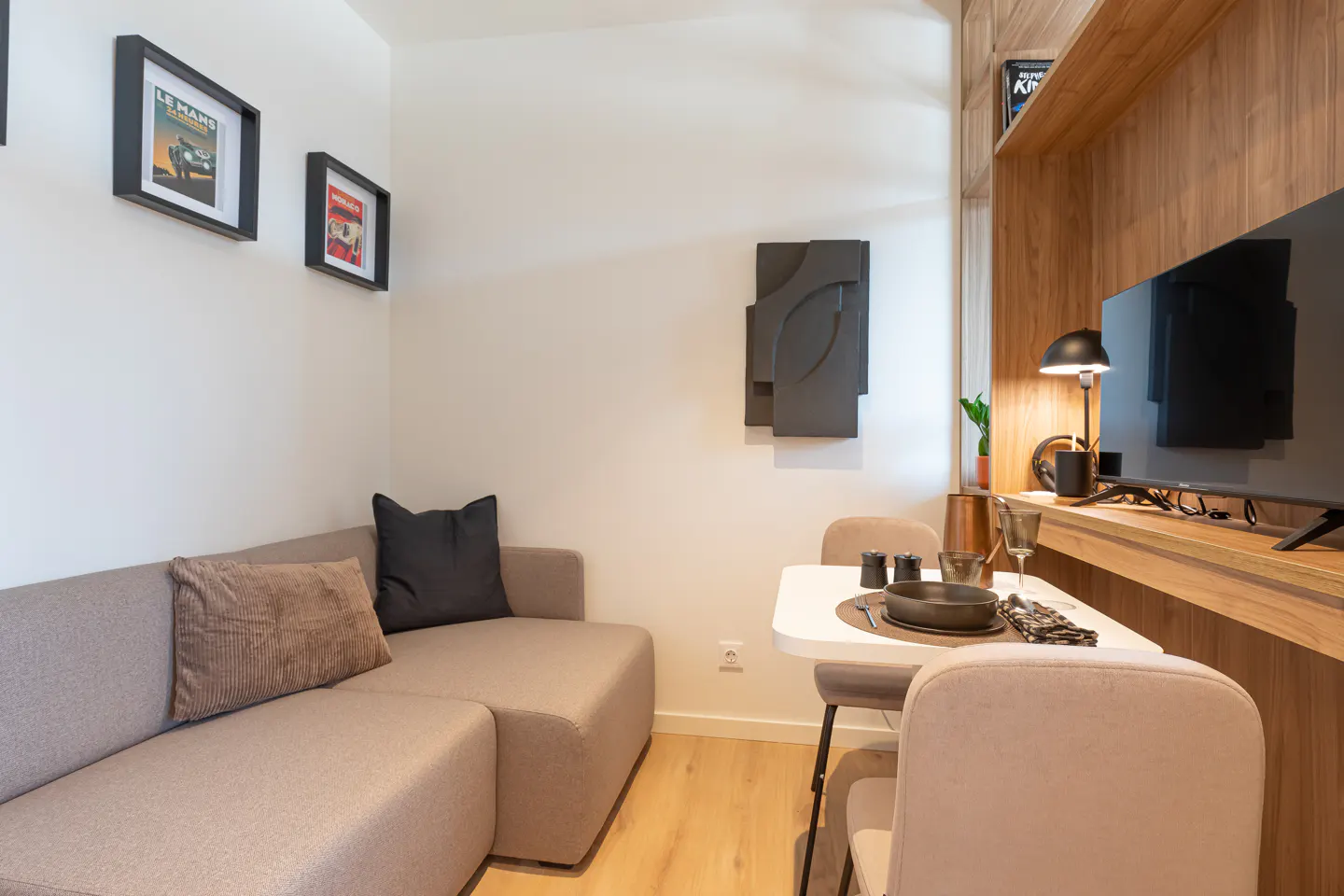 Living room with a beige sofa, white dining table set for two, and a wooden entertainment center with a TV. Artwork adorns the white walls.
