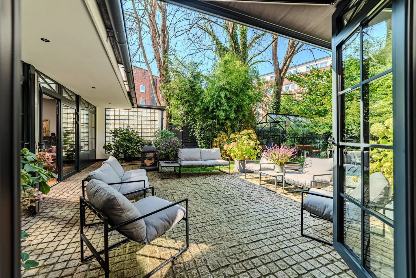 A patio with gray cushioned seating and black frames, a brick floor, and a garden view through open black-framed doors.