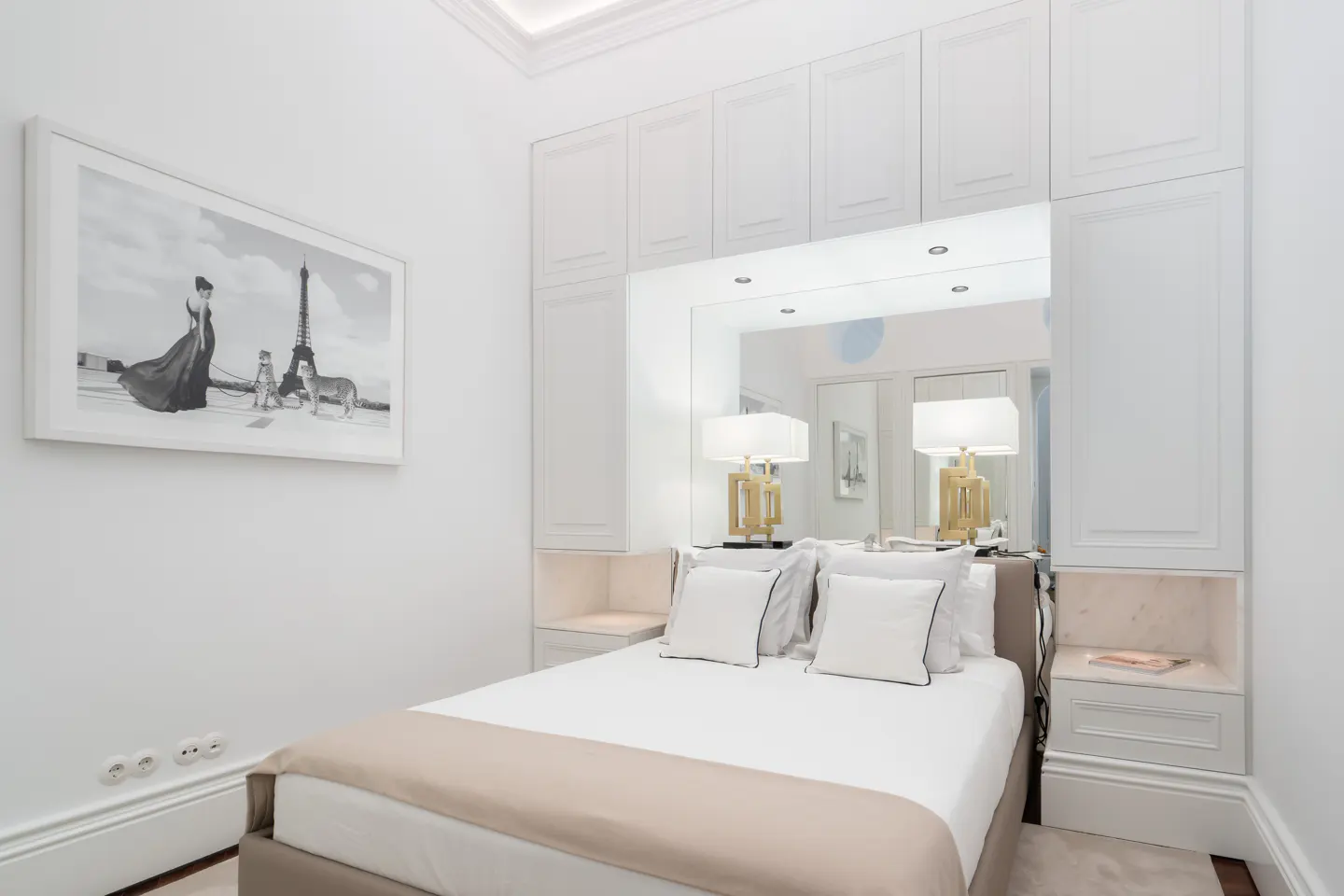Bright bedroom with white walls, built-in cabinets, and a bed with white linens and a tan throw. A framed black and white print hangs above the bed.