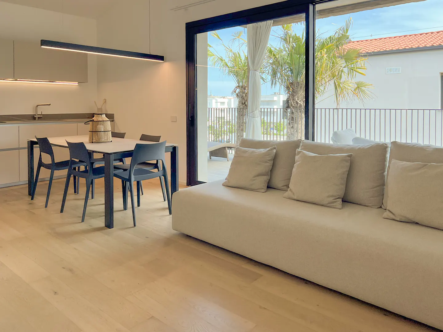 Modern open-plan living area with a beige sofa, dining table with dark chairs, and a balcony view with palm trees.