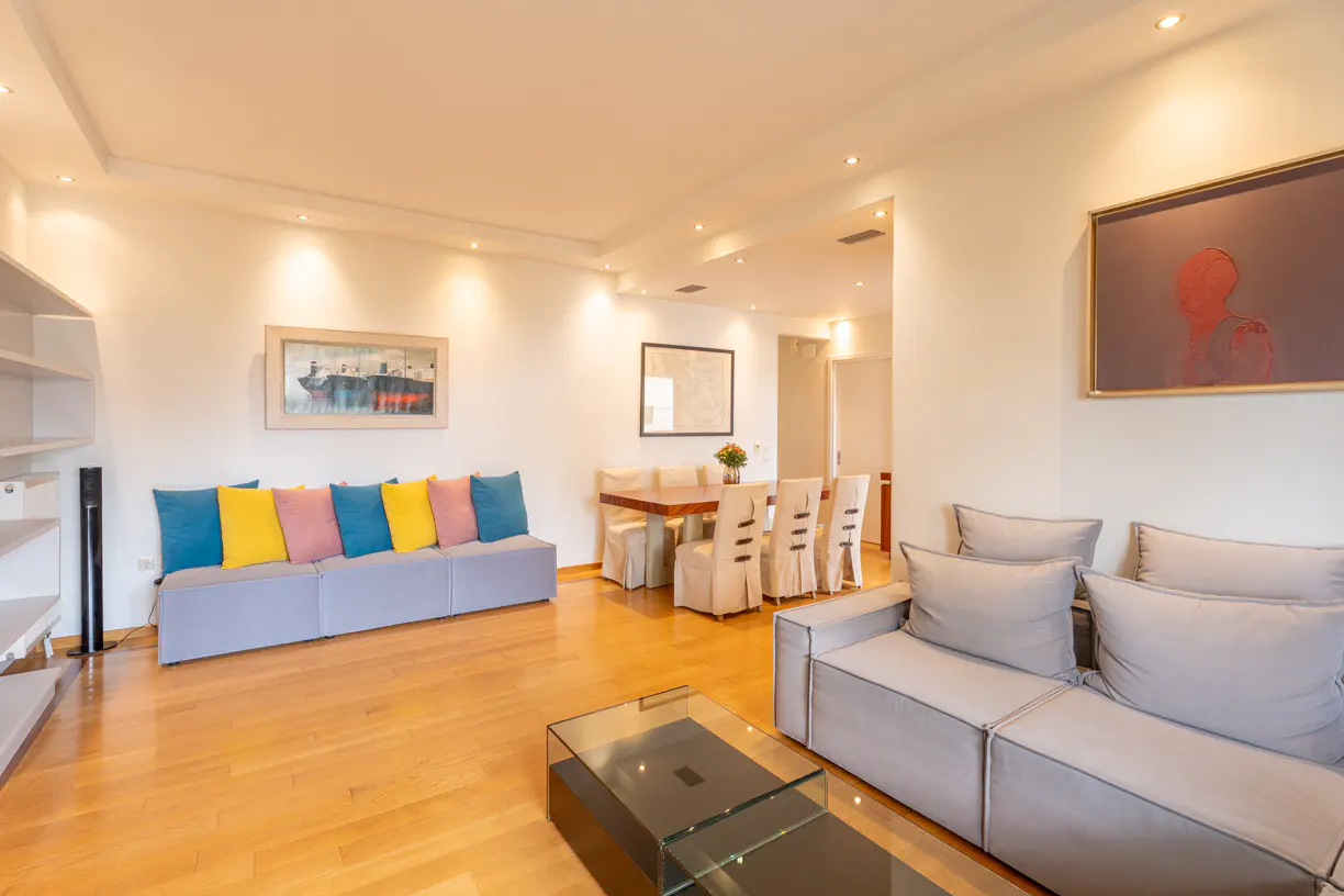 Bright living room with wood floors, gray sofas, and colorful pillows. A dining table and artwork adorn the walls.