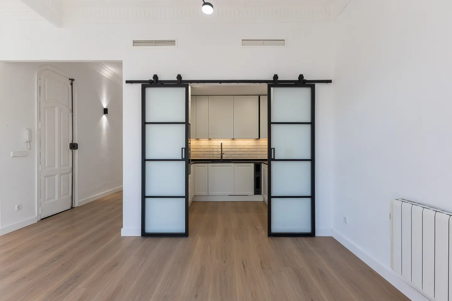 A bright, modern apartment with wood floors and white walls. Black framed sliding doors open to a white kitchen. A white door is visible in the background.