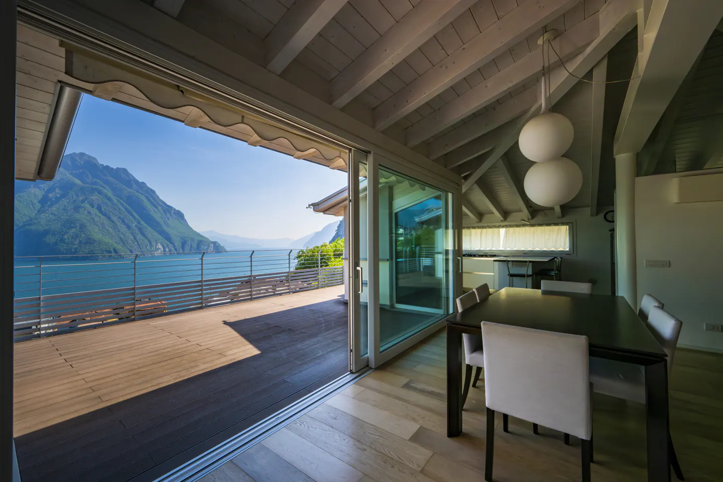 Interior view of a dining room with a dark wood table and white chairs, open sliding doors lead to a deck with a lake and mountain view.