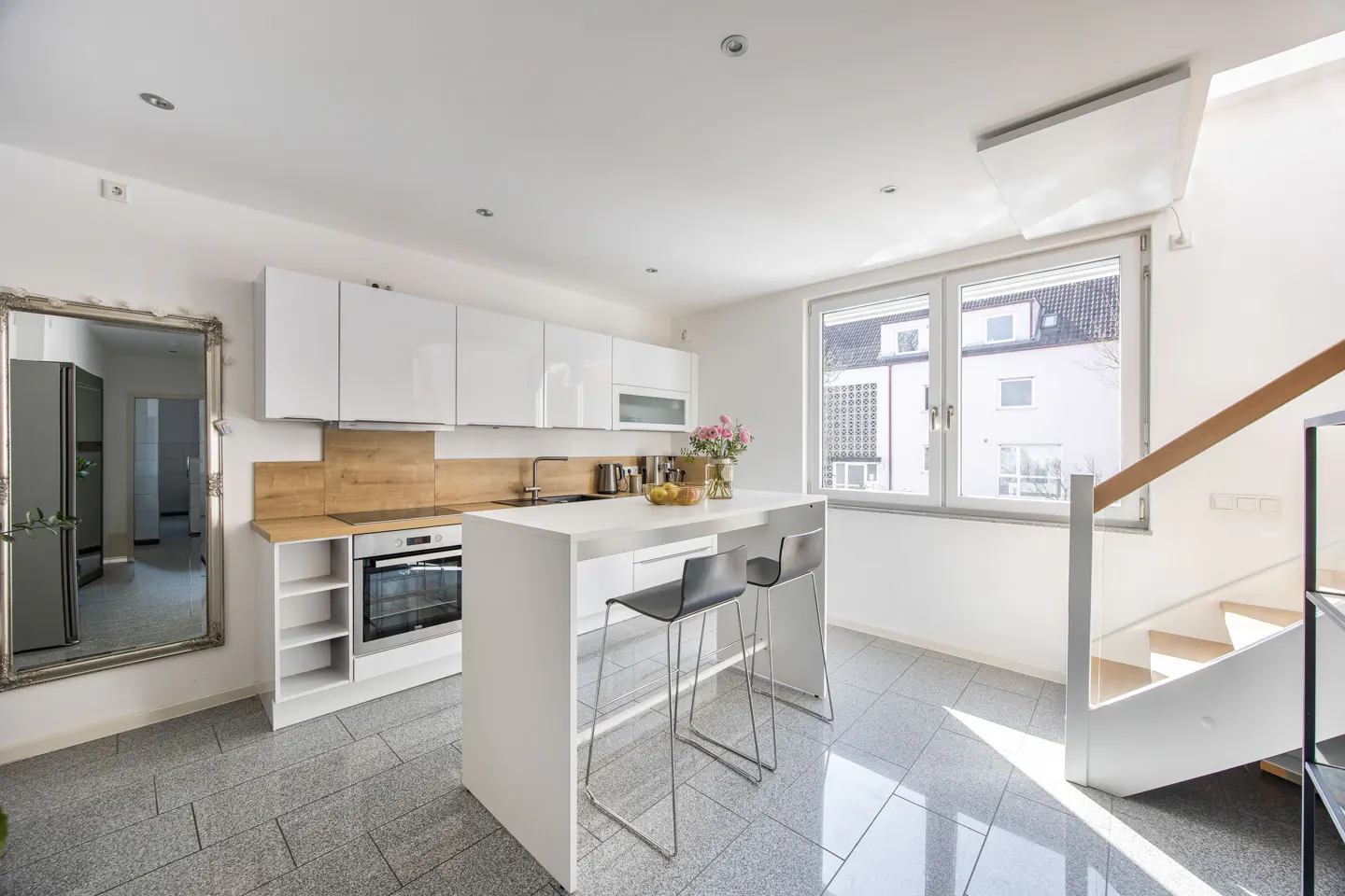 Bright kitchen with white cabinets, island with two stools, and a staircase with glass railing. Gray tile floor and a large mirror.