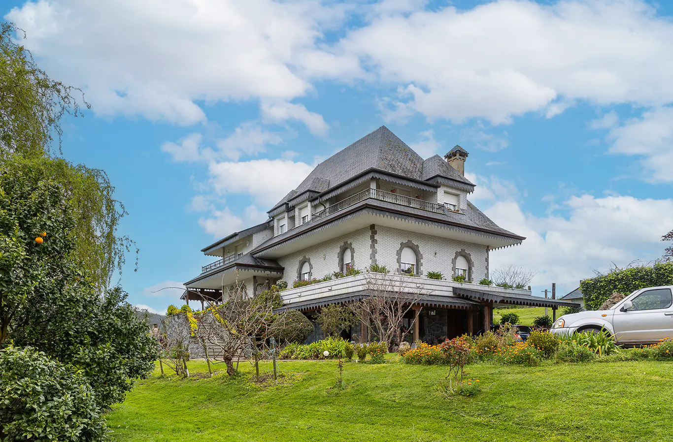 A large, white, multi-story house with a gray roof sits on a green lawn under a blue, cloudy sky.
