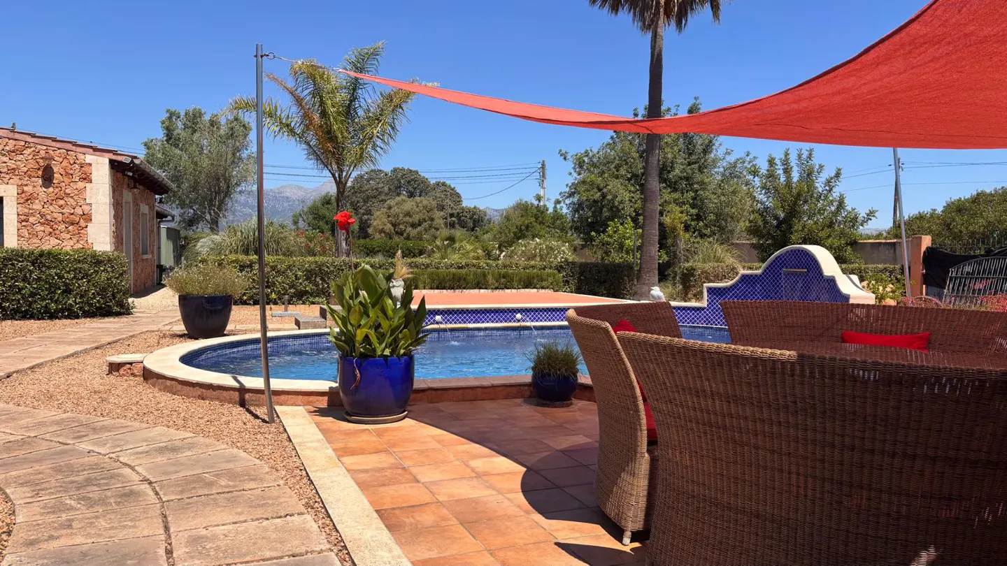 Outdoor patio with a pool, wicker furniture, and a red sunshade. Blue potted plants add color to the scene.