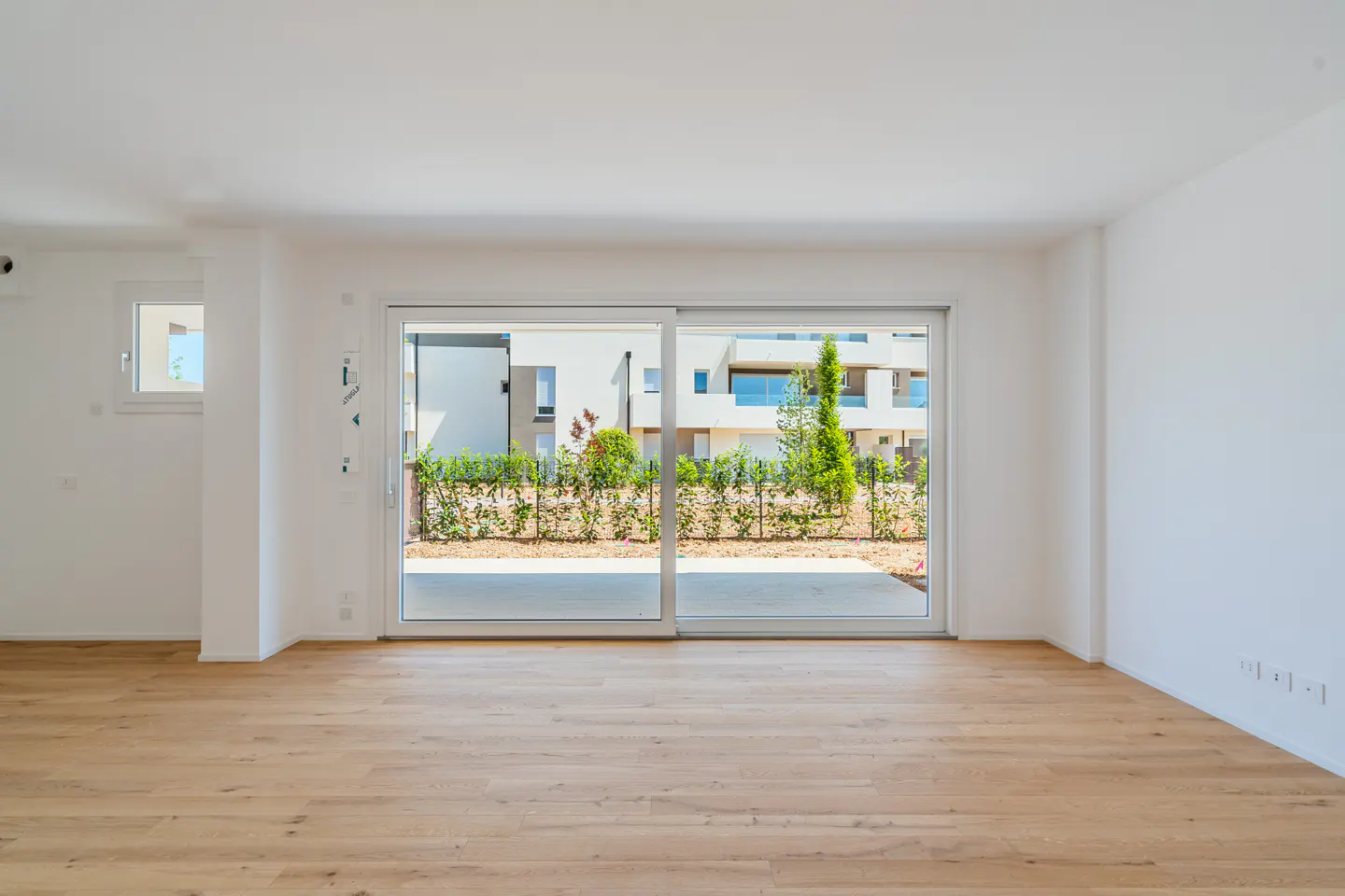 Empty room with light wood floors and white walls. Sliding glass doors open to a patio and garden.