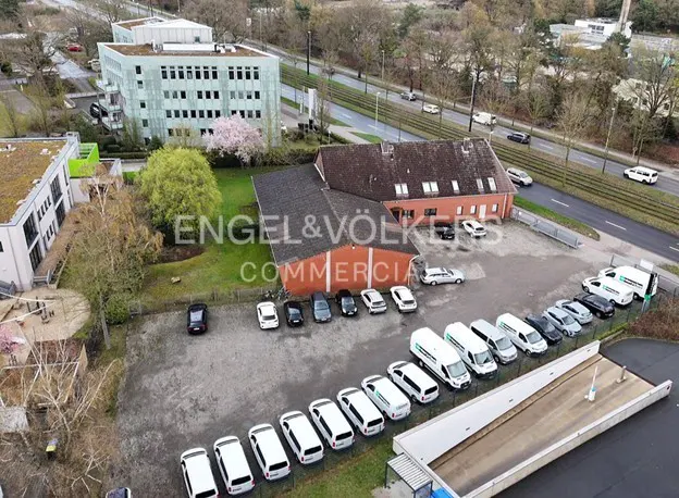 Aerial view of a commercial property with a red brick building, parking lot filled with white vans, and a modern office building nearby.