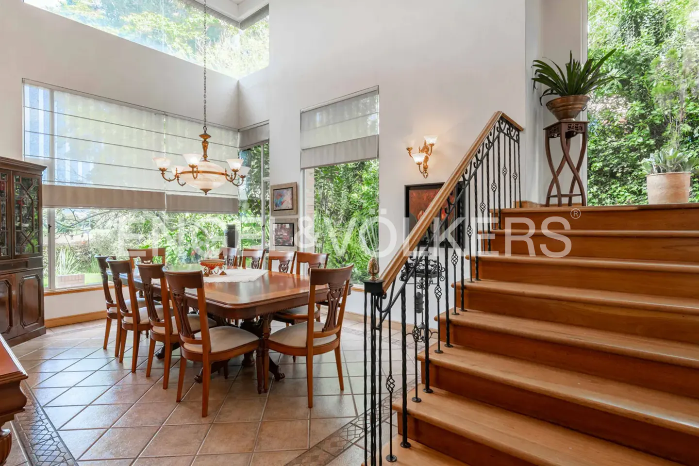 Bright dining room with a wood table, chairs, and chandelier. Large windows overlook greenery. A wooden staircase with black iron railing is on the right.