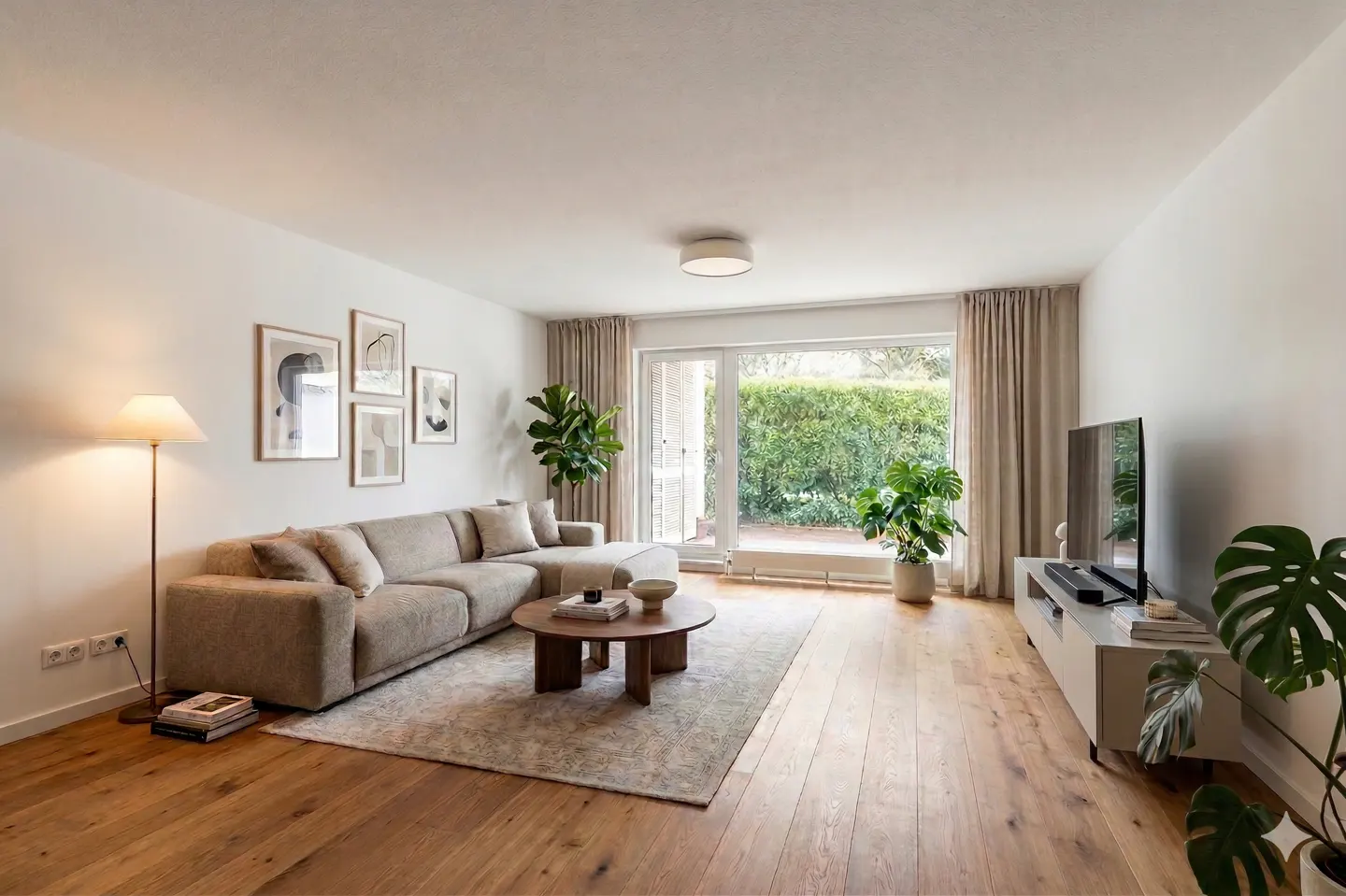 Bright living room with hardwood floors, a beige sectional sofa, round coffee table, and a TV. Artwork adorns the walls. Plants add a touch of nature.