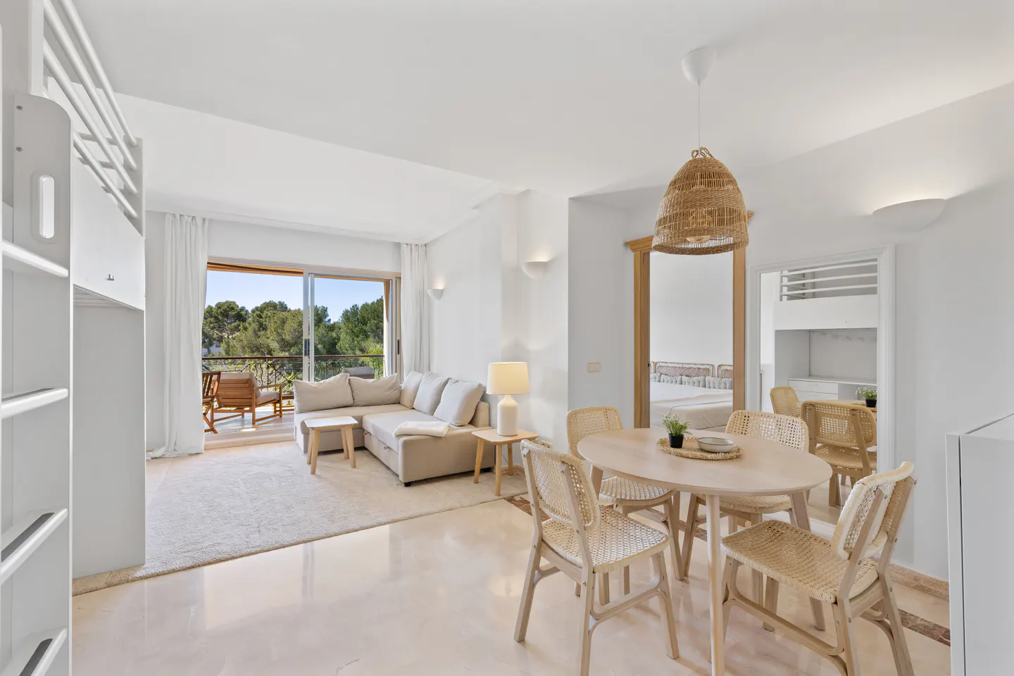 Bright living space with a beige sofa, round table with wicker chairs, and a balcony overlooking trees.