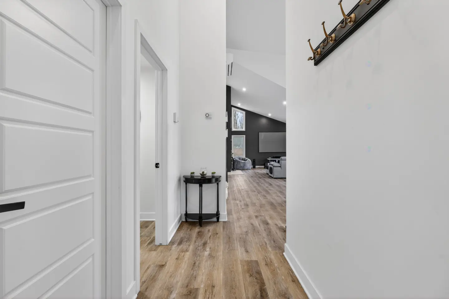 Hallway view with white walls, light wood floors, and a black coat rack. A black table sits near an open doorway leading to a living room.
