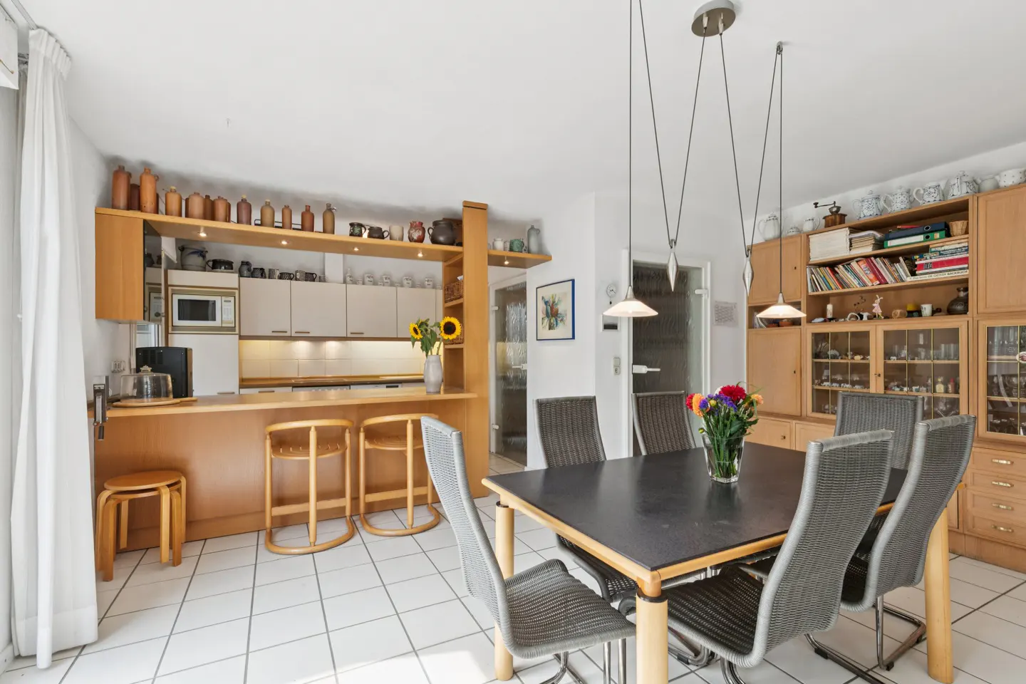 A bright kitchen and dining area with white tile floors, light wood cabinets, and a black dining table with six gray chairs.