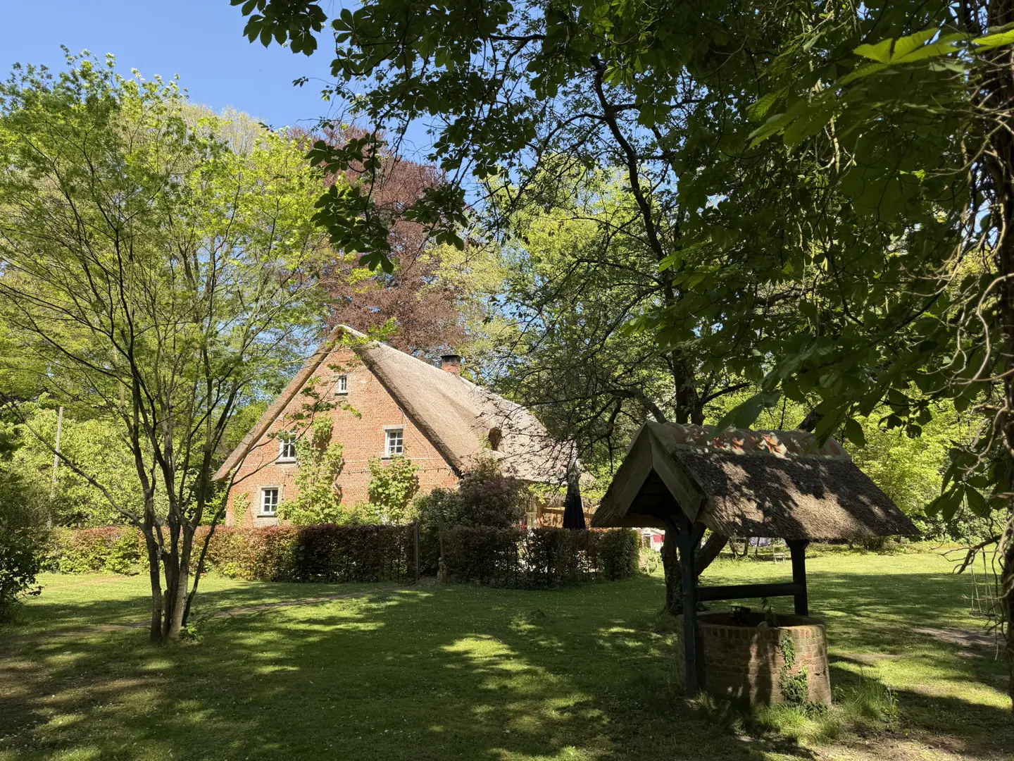 Exterior view of a red brick house with a thatched roof, surrounded by green trees and a well in the foreground.