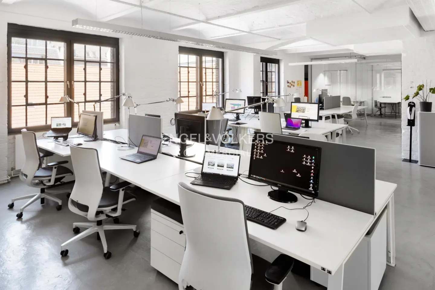 Bright, modern office space with white desks, chairs, and computers. Large windows provide natural light. Gray dividers offer privacy.