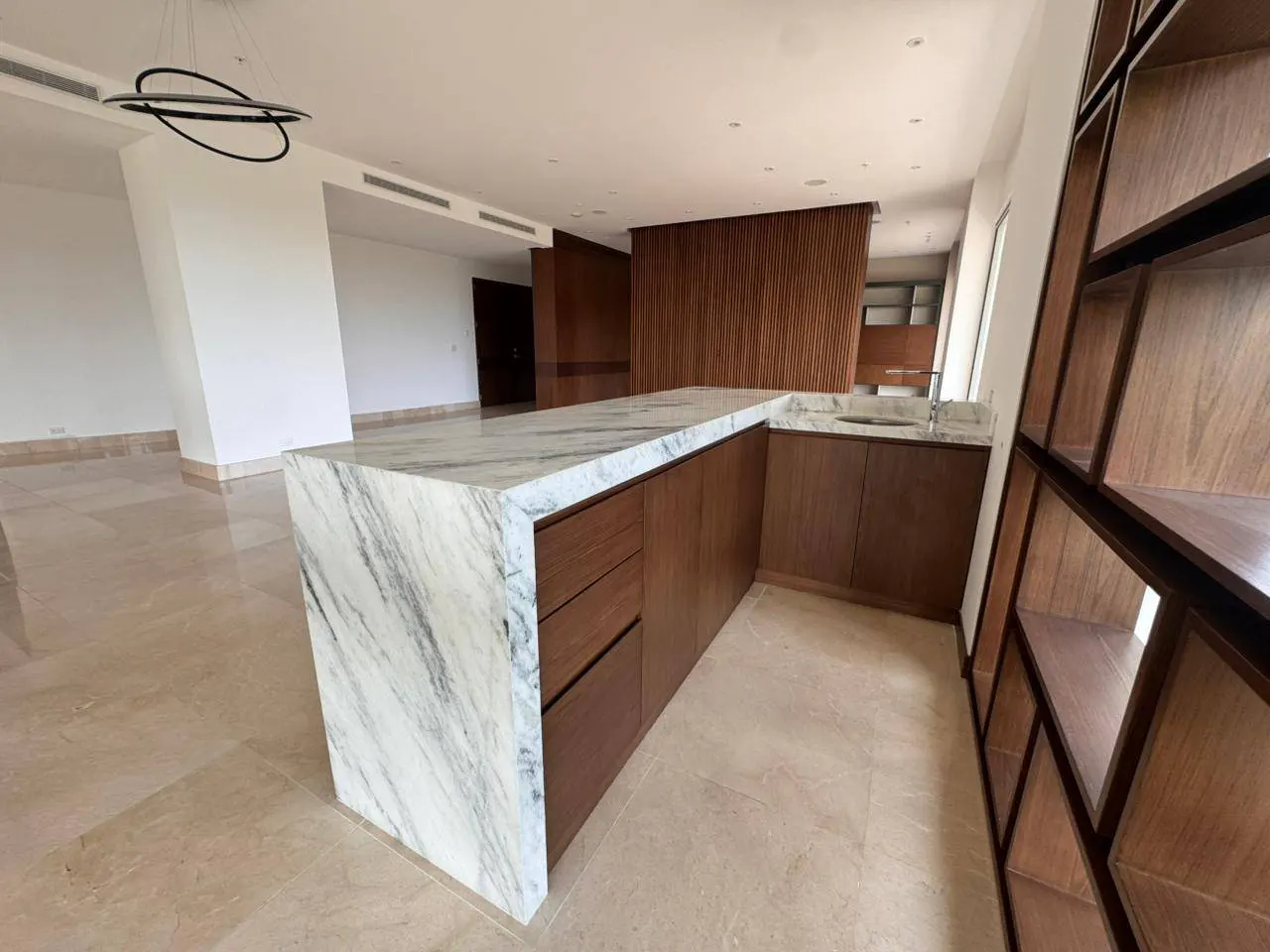 A modern kitchen with a marble countertop island and wooden cabinets. A wooden bookshelf is on the right.