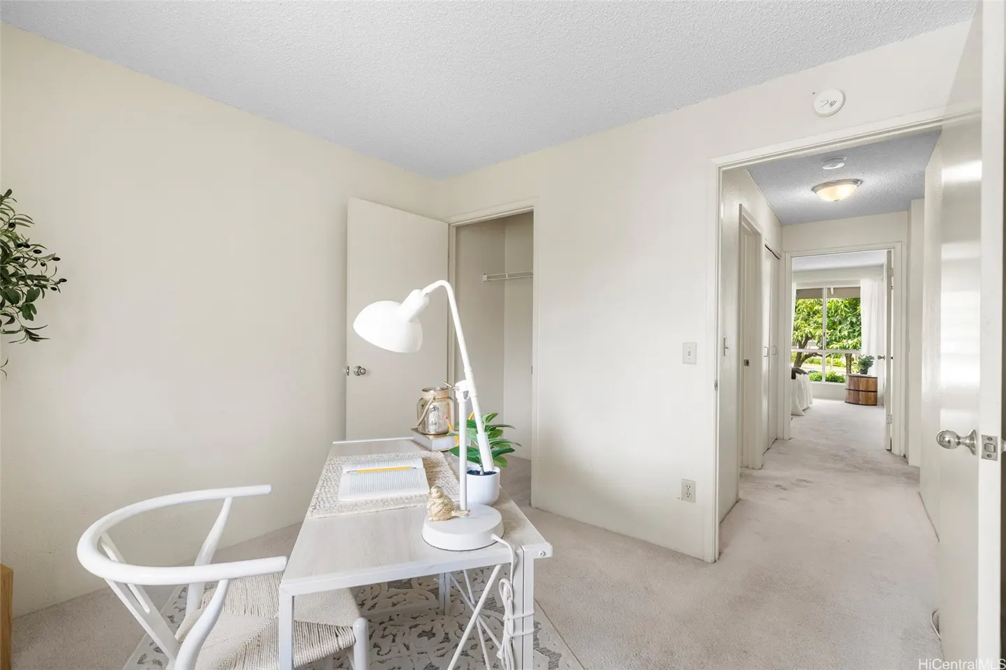 A bright home office with a white desk, lamp, and chair, featuring an open closet and a view into a hallway.