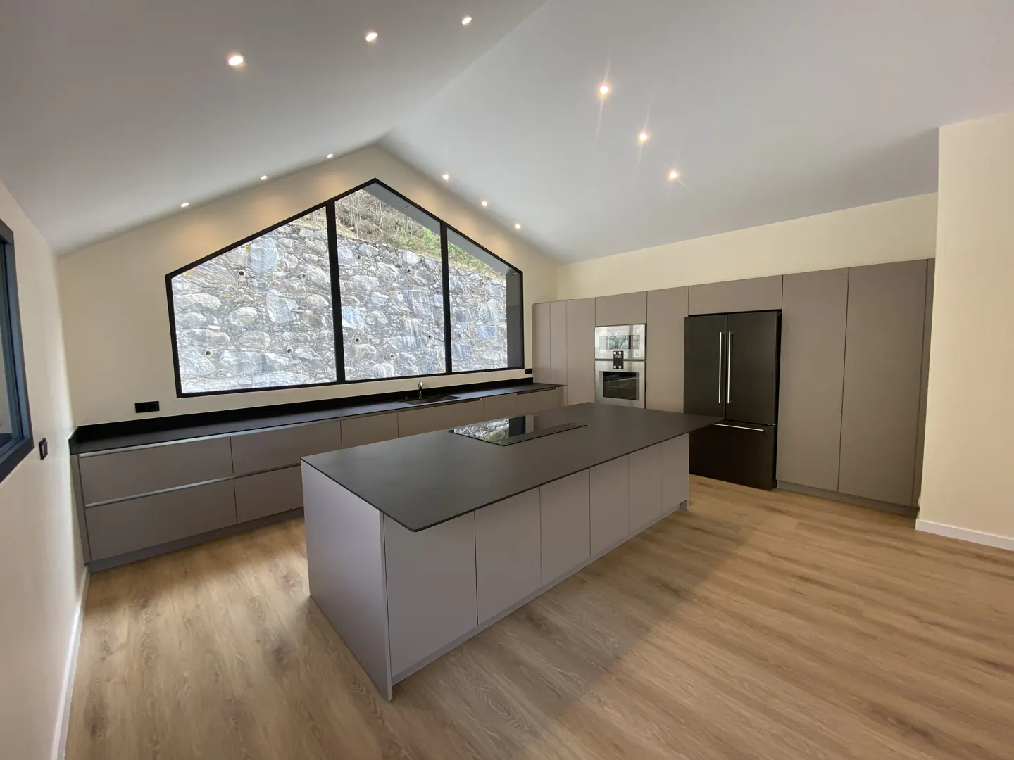 Modern kitchen with light wood floors, gray cabinets, black countertops, and a large window with a view of a stone wall.