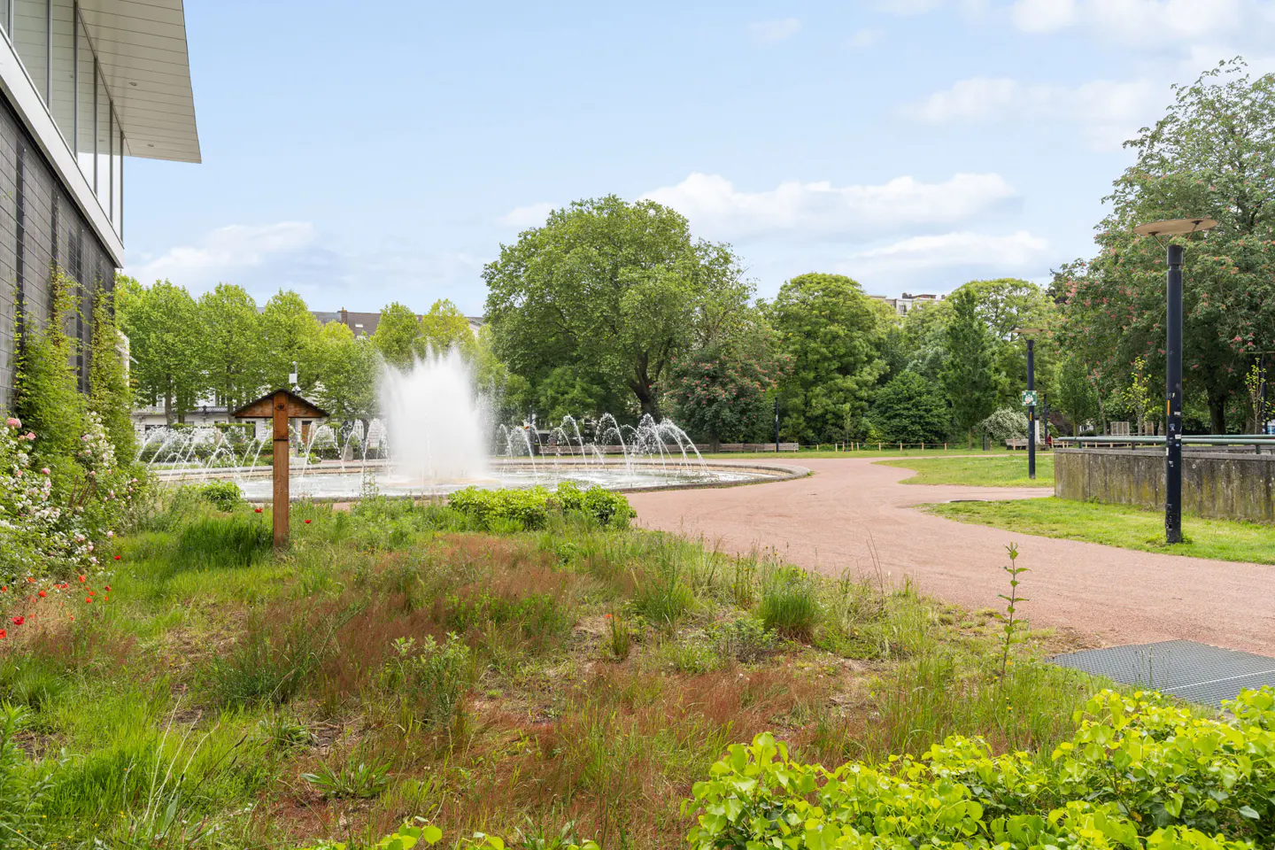 A park with a large fountain in the center, surrounded by green grass and trees. A red gravel path winds through the park.