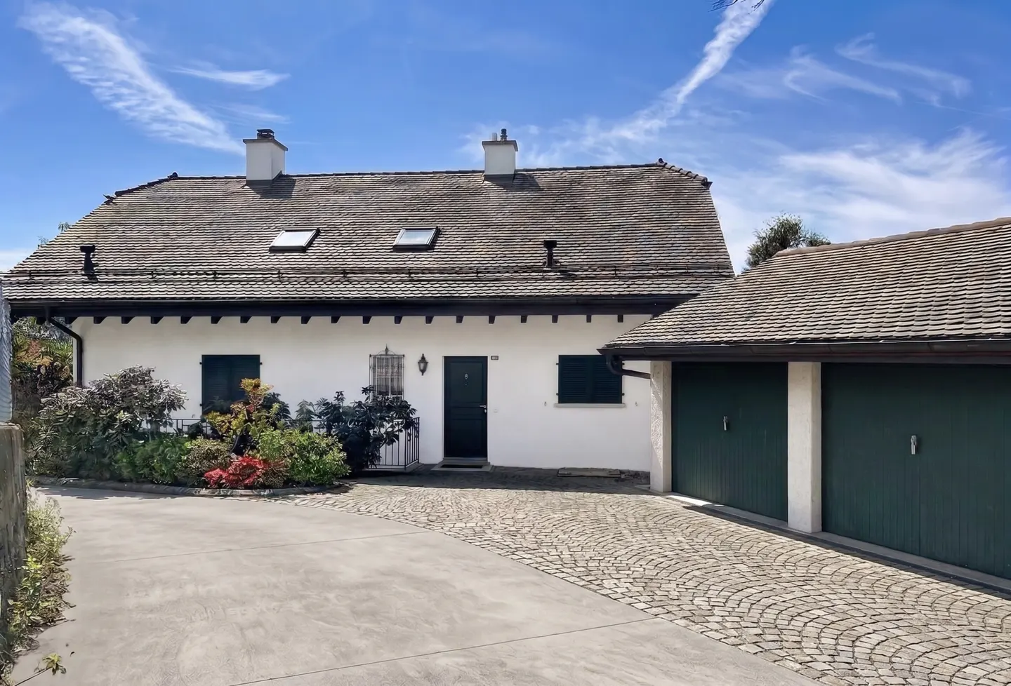 White house with a gray roof, dark green shutters and garage doors, and a cobblestone driveway under a blue sky.