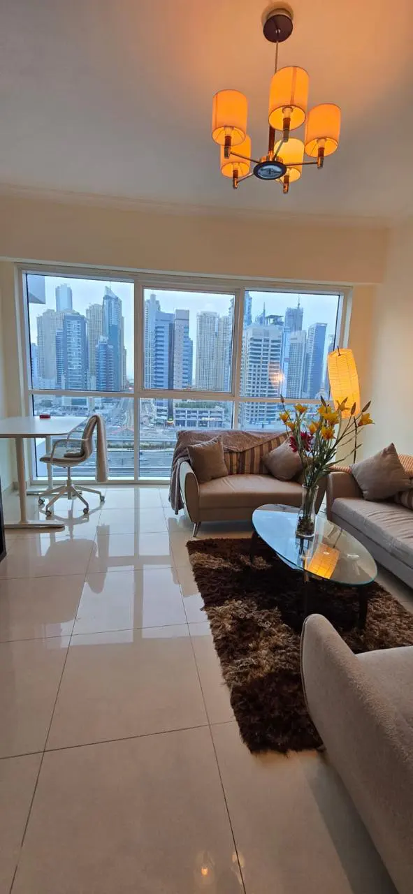 Living room with city view. Beige sofas, glass table with flowers, brown rug, and desk by the window.