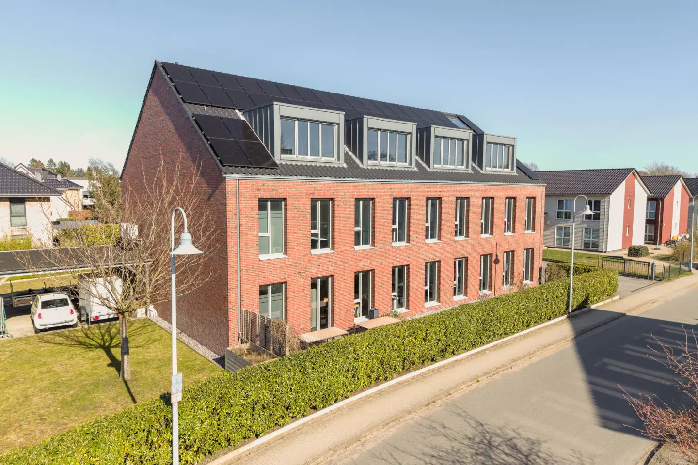 Modern red brick apartment building with solar panels on the roof, surrounded by a green hedge and a paved street.