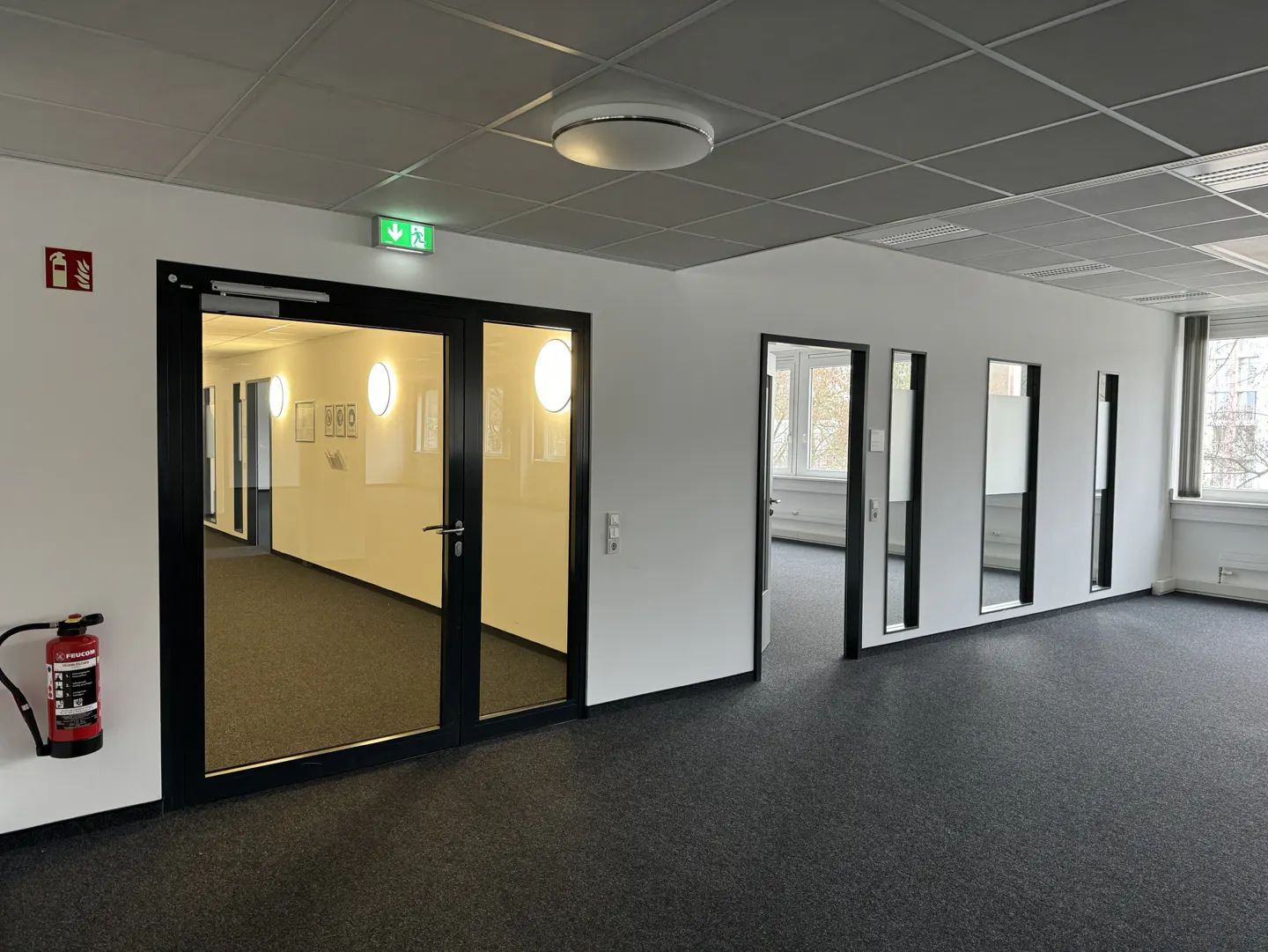 Empty office space with gray carpet, white walls, and black door frames. A fire extinguisher is mounted near a glass door.
