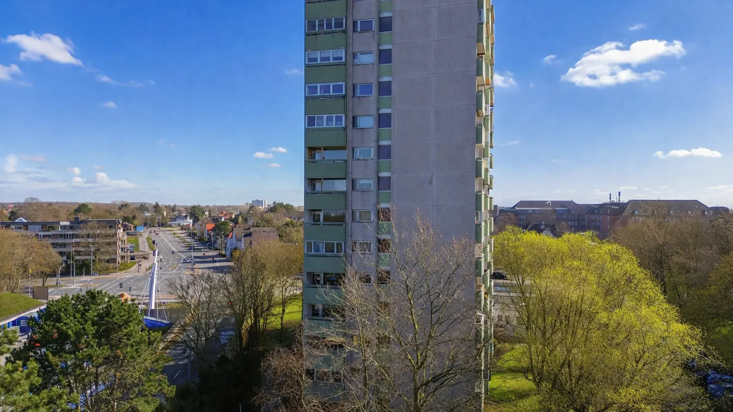 Exterior view of a tall, gray apartment building with green accents, set against a blue sky and a cityscape background.