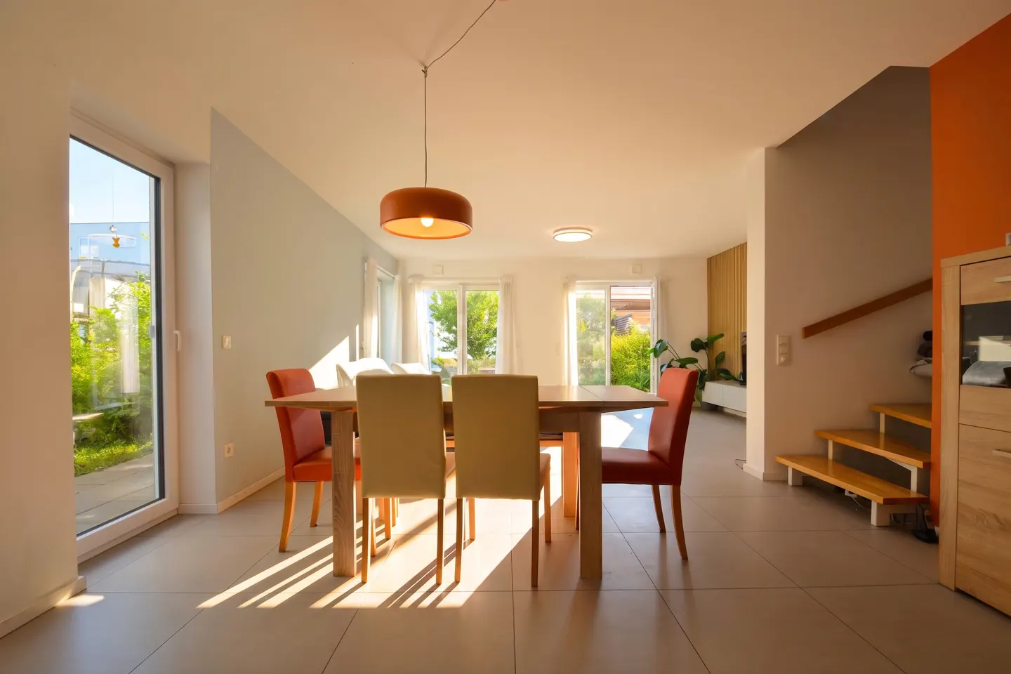 Bright dining room with a wood table, orange and beige chairs, and a modern orange pendant light. Sunlight streams through the windows.