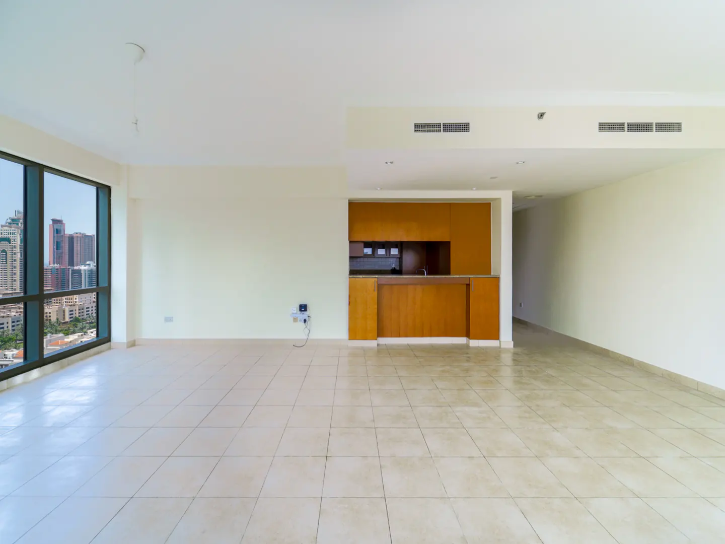 An empty, bright room with beige tile floors, white walls, and a kitchen area with wood cabinets. A large window shows a city view.