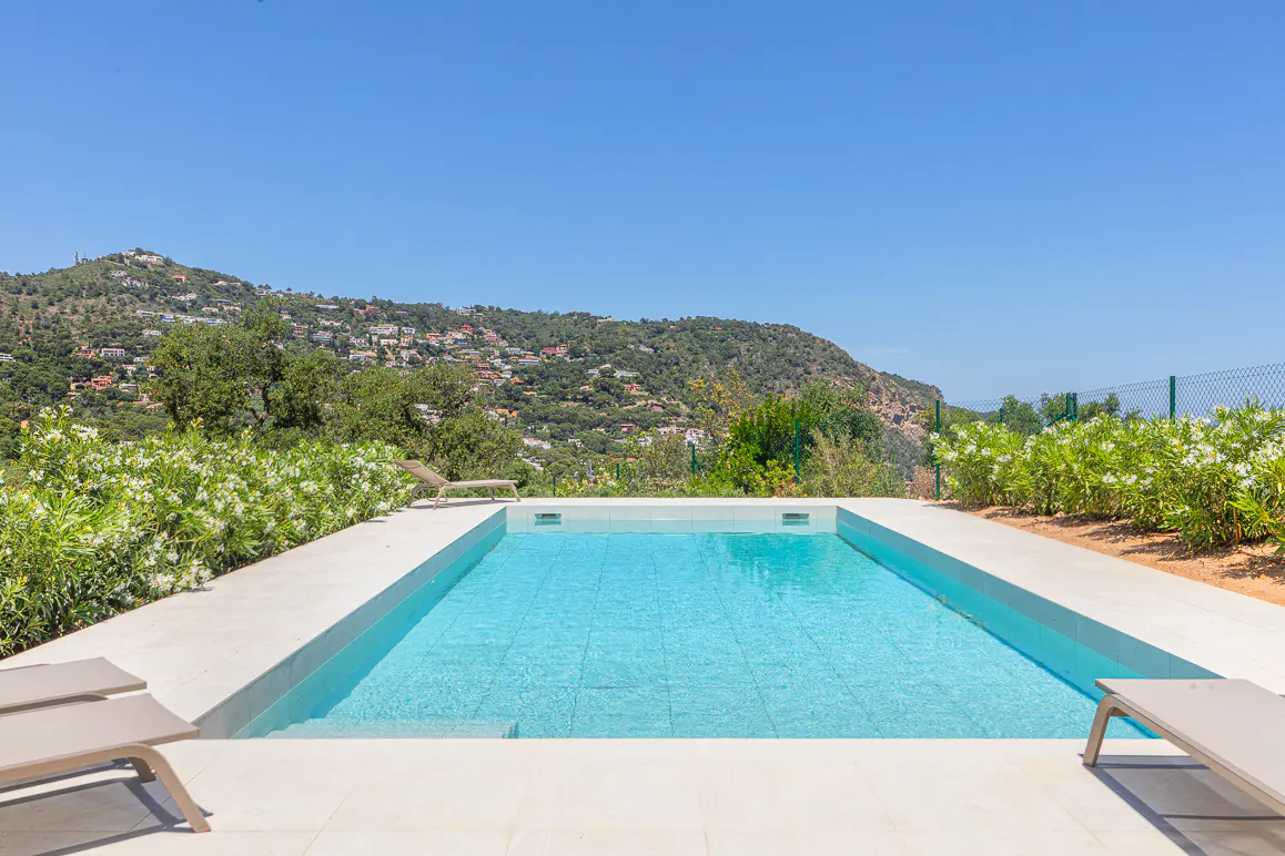 Rectangular swimming pool with light blue water, surrounded by white stone decking, lounge chairs, and a mountain view.
