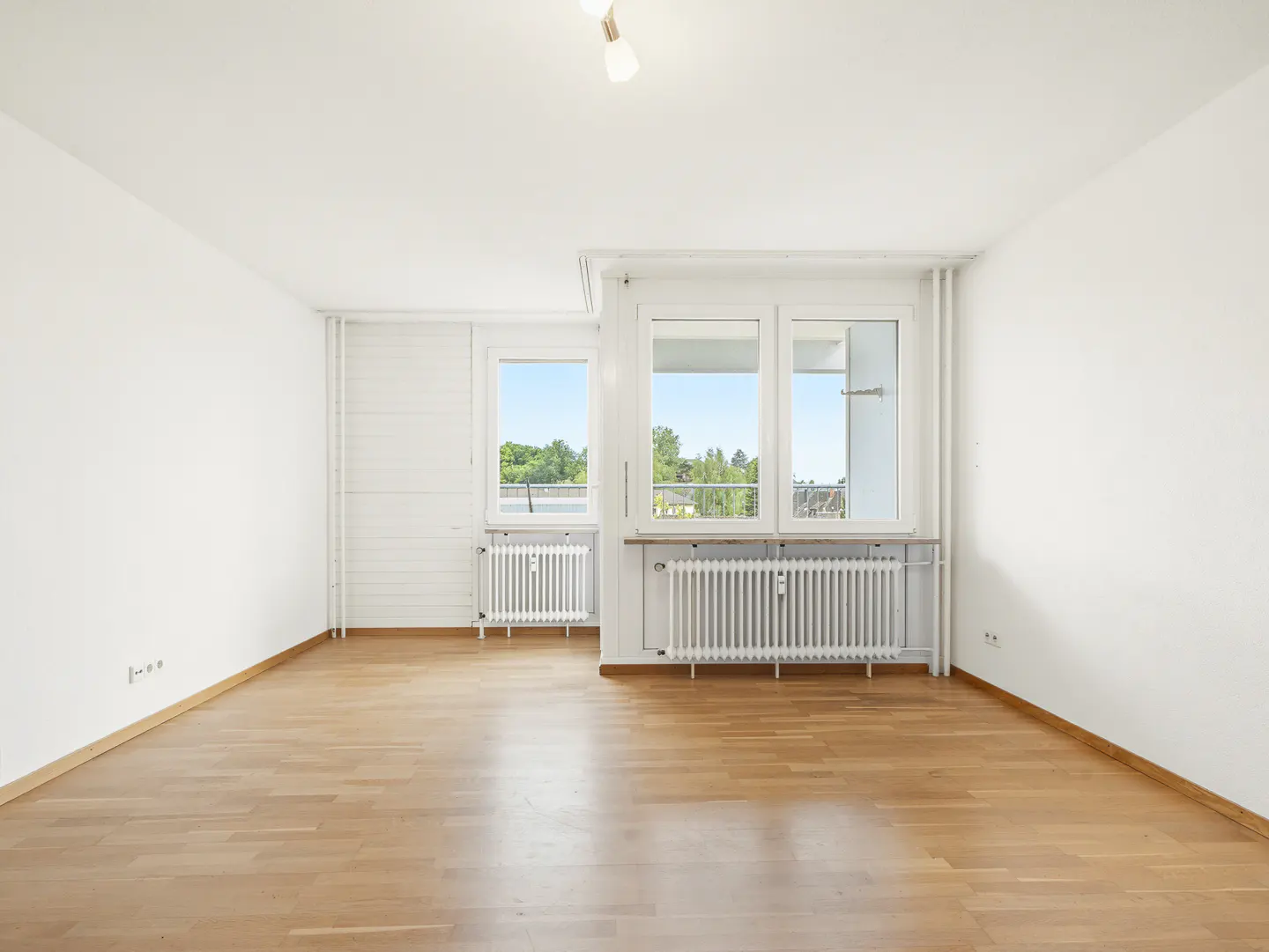 Bright, empty room with hardwood floors, white walls, two windows with white frames, and white radiators below them.