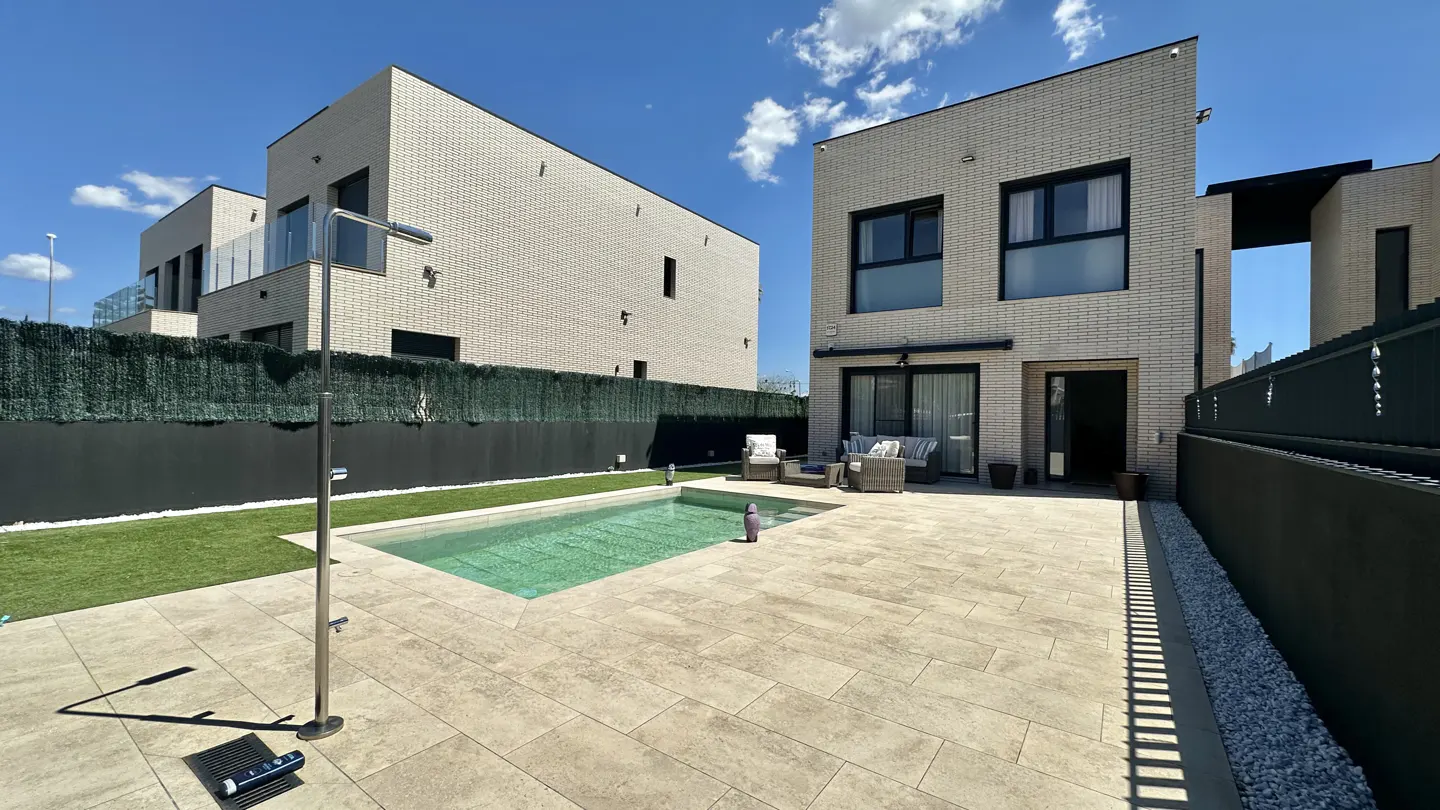 Modern home exterior with a pool, patio furniture, and a shower. The house is light brick with black trim and a green hedge.