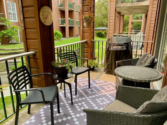 A cozy outdoor patio with black chairs, a wicker table and chairs, and a Coleman grill. A gray and white patterned rug covers the floor.