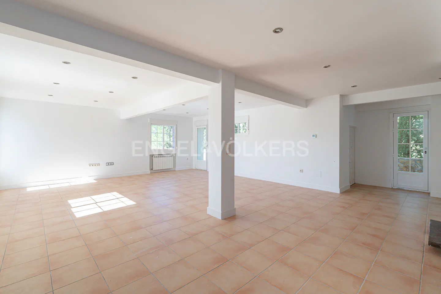 Bright, empty room with white walls, terracotta tile floor, and support column. Windows and a glass-paneled door let in natural light.