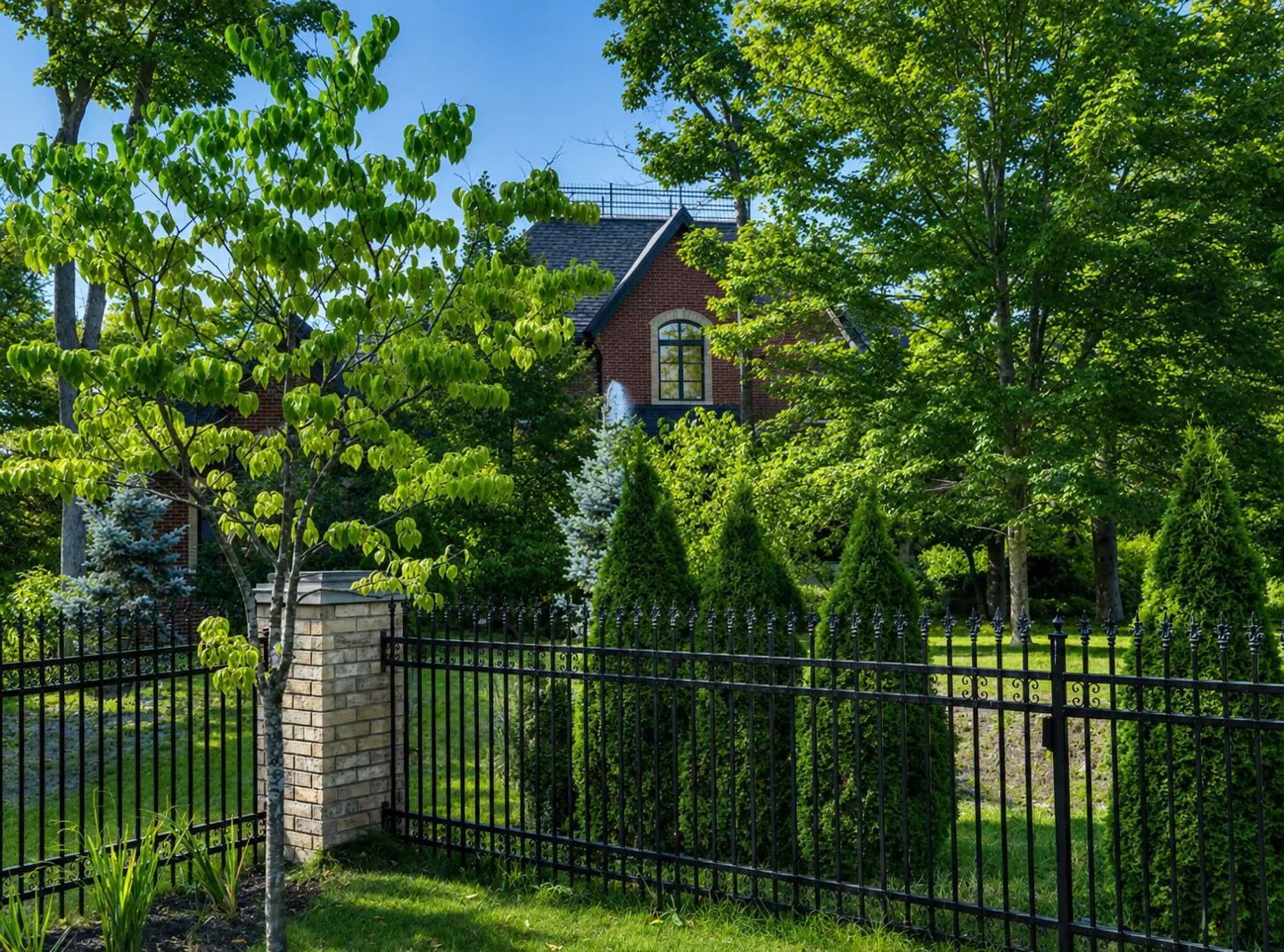 A brick house is partially obscured by green trees and bushes behind a black metal fence.