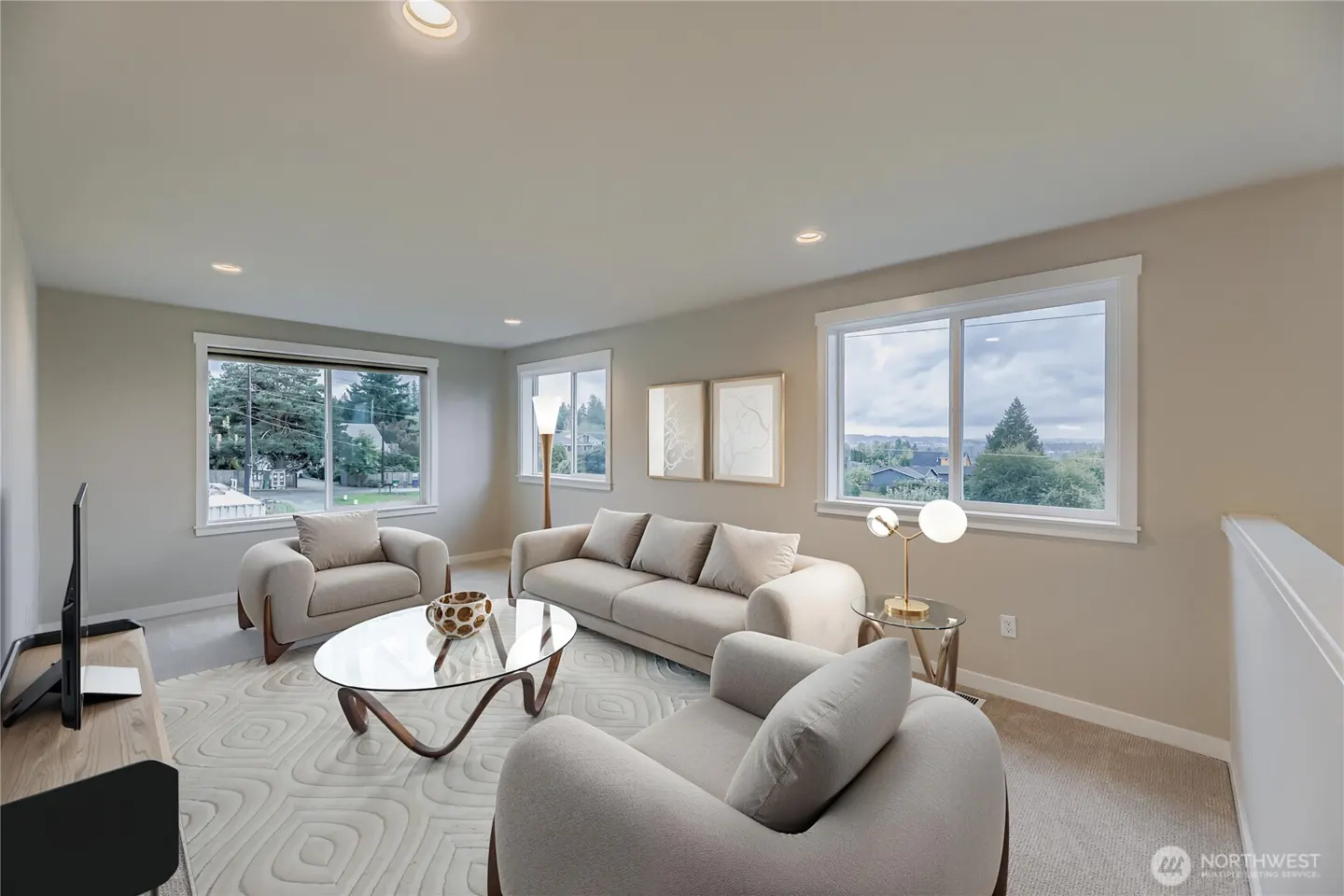Bright living room with beige furniture, a glass coffee table, patterned rug, and large windows overlooking greenery.