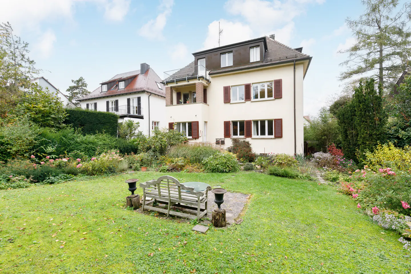 Exterior view of a two-story cream house with brown shutters and a green lawn with a bench.