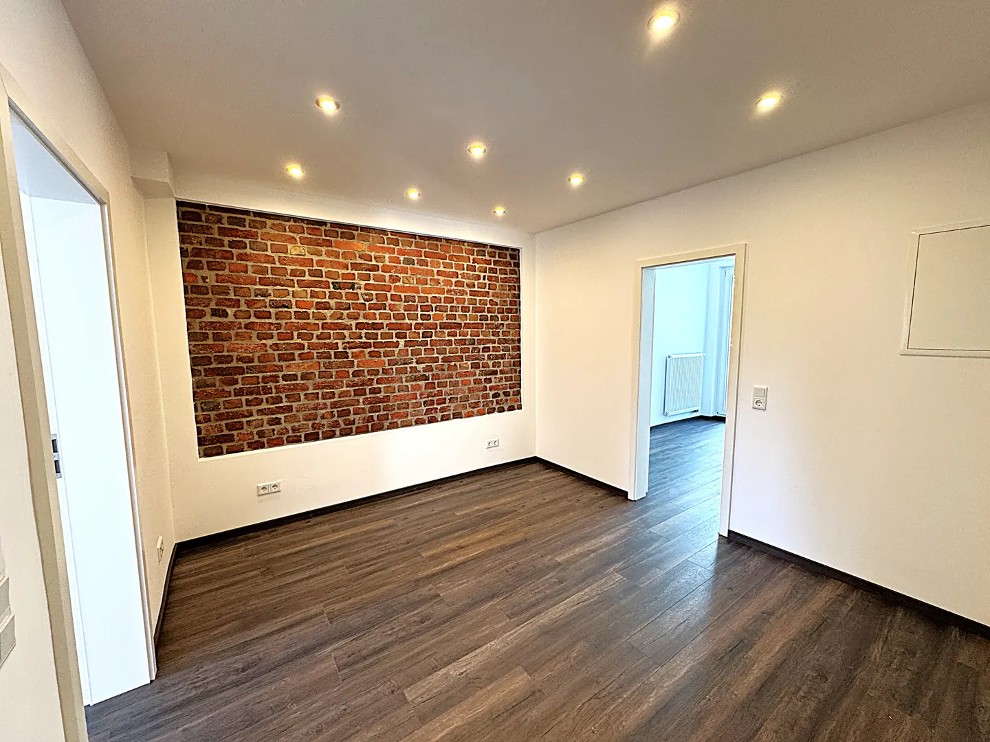 An empty room with dark wood floors, white walls, and a red brick accent wall. Recessed lighting illuminates the space. A doorway leads to another room.