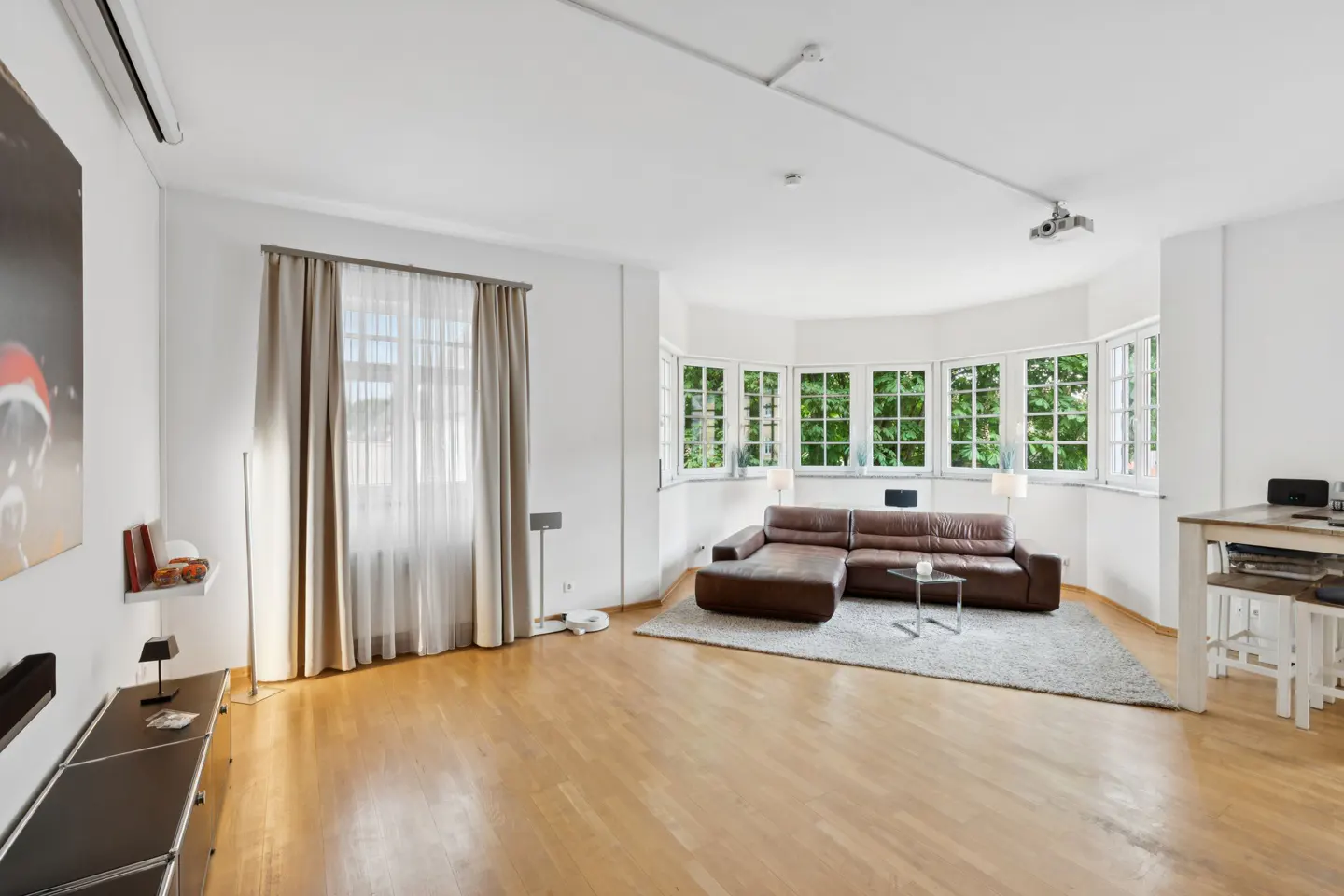 Bright living room with hardwood floors, a brown leather sectional on a grey rug, and large bay windows with beige curtains.