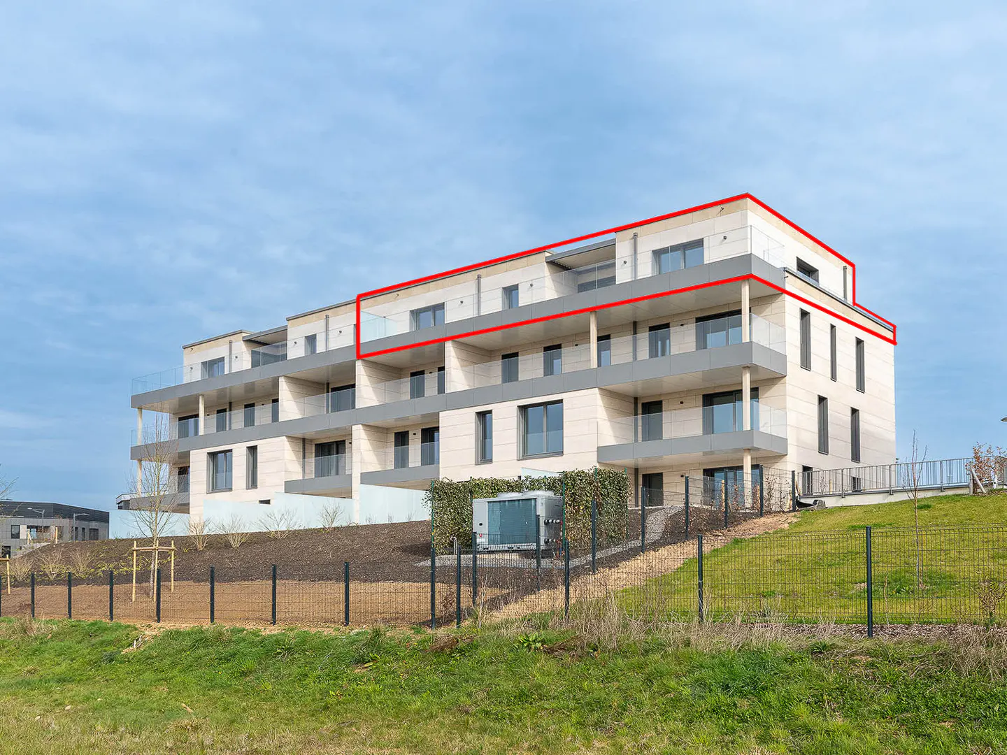 Modern apartment building with balconies, outlined in red, against a blue sky and green grass.
