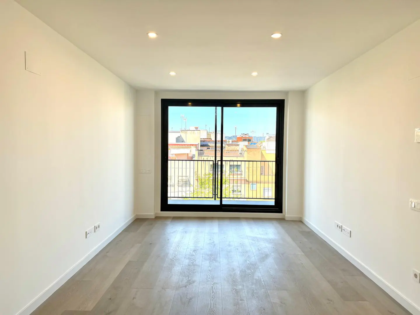 Bright, empty room with white walls, gray wood floor, and black-framed sliding glass doors to a balcony with city views.