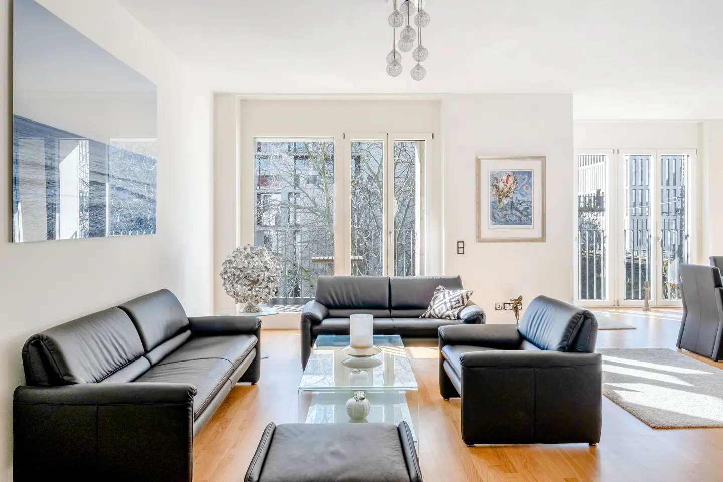 Bright living room with black leather sofas, glass tables, and wood floors. Large windows offer a view of trees.