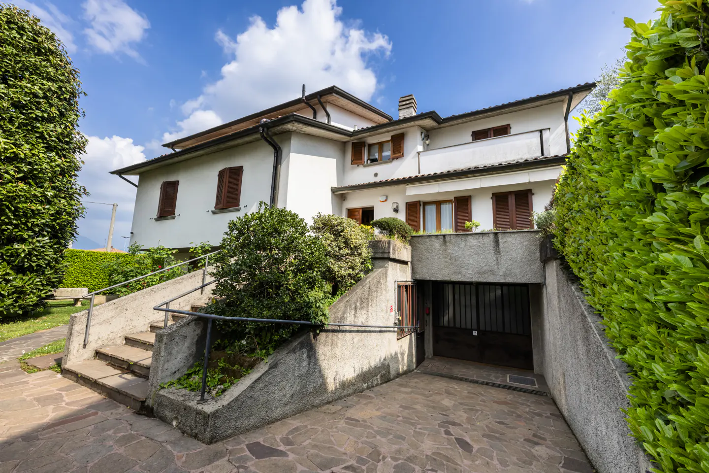 A two-story white house with brown shutters and a garage entrance on the ground floor. Steps lead up to the main level. Green bushes surround the house.