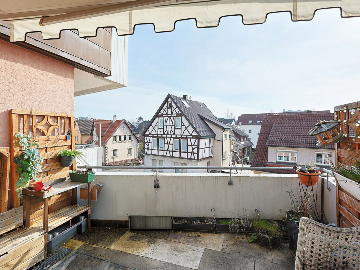 Balcony view with potted plants, wooden furniture, and a view of houses with red roofs under a bright sky.