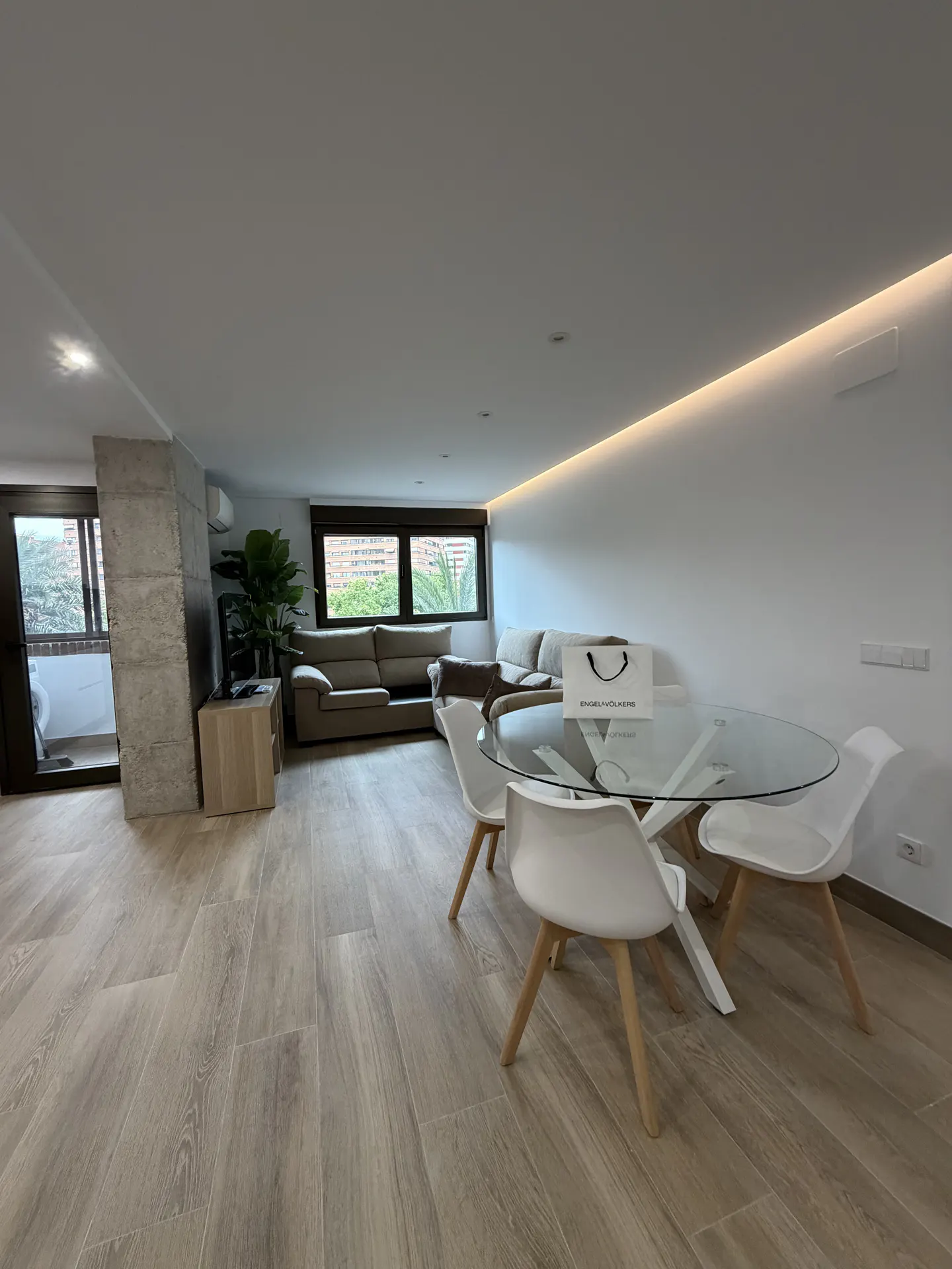 Bright, modern living room with a glass table, white chairs, and a beige sofa. A shopping bag sits on the table. Wood-look flooring.