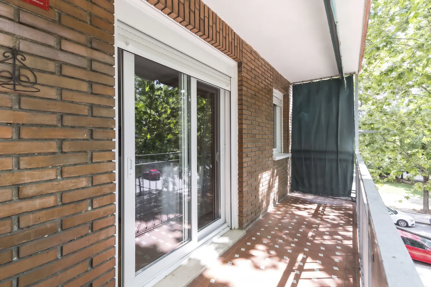 A balcony with red brick walls, a white sliding glass door, and red tiled floor. A green curtain hangs on the right side.