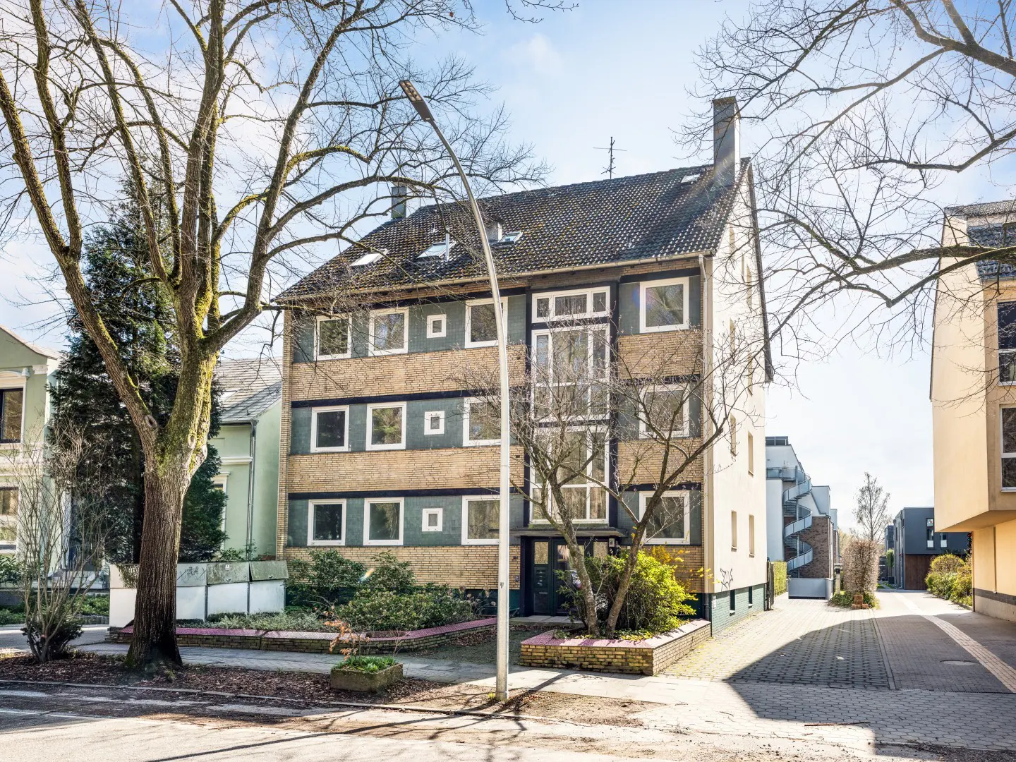 A three-story apartment building with a gray roof and a mix of tan and green walls. Bare trees frame the building.