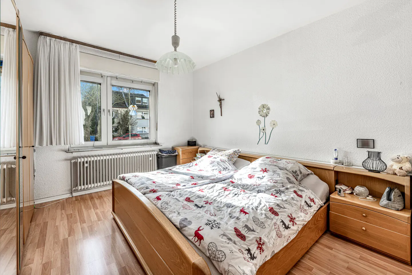Bright bedroom with a wooden bed, patterned duvet, and bedside tables. A window with curtains is on the left, and wall decorations are on the right.